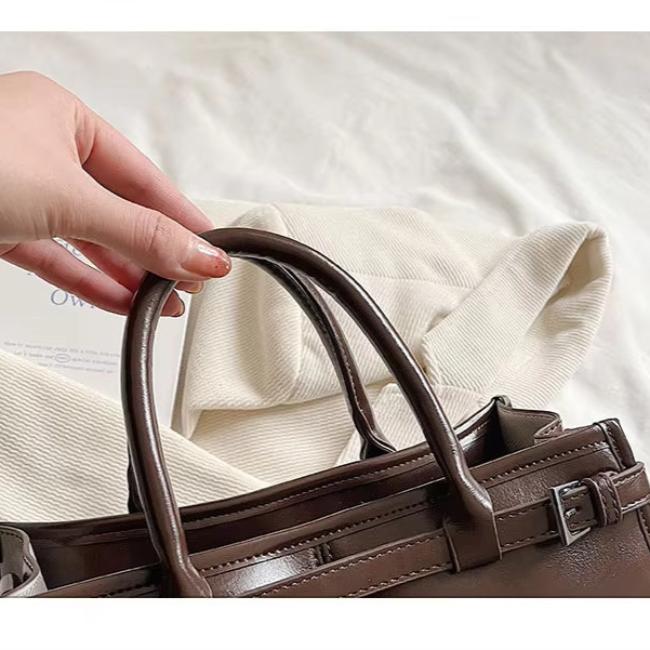 Borsa a mano in pelle marrone lucida con manici, su sfondo di tessuto bianco. Accessorio moda elegante, ideale per shopping e lavoro.