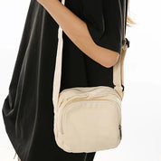 Borsa a tracolla beige in tessuto, indossata da una persona con abito nero. Design compatto, ideale per moda casual e uso quotidiano.