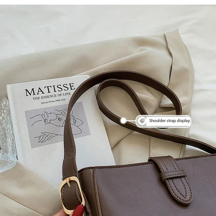 Borsa a tracolla marrone su rivista d'arte Matisse, con fibbia dorata, su sfondo beige. Accessorio moda elegante e versatile.