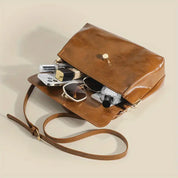 Borsa a tracolla in pelle marrone aperta, contenente occhiali da sole, rossetto, specchietto e crema. Accessorio moda elegante e versatile.