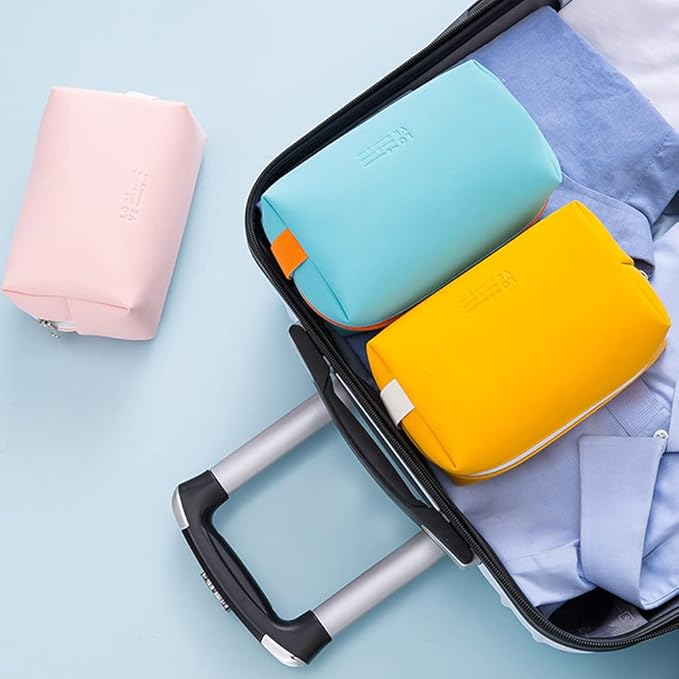 Valigia aperta con due beauty case colorati, uno azzurro e uno giallo, su camicia blu. Beauty case rosa accanto. Organizzazione viaggio.