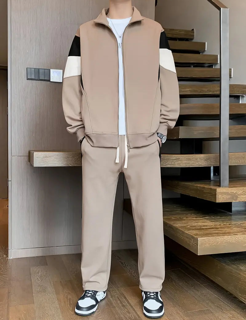 Tuta sportiva beige da uomo con giacca a zip e pantaloni abbinati, dettagli bianchi e neri, indossata con sneakers bianche e nere su pavimento in legno.