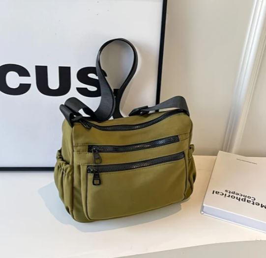 Borsa a tracolla verde oliva con tasche multiple e cerniere nere, posizionata su un tavolo bianco accanto a un libro e una cornice.