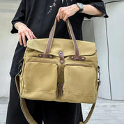 Borsa a tracolla in tela beige con manici in pelle marrone, tasche frontali con cerniera, indossata da una persona in abbigliamento casual.