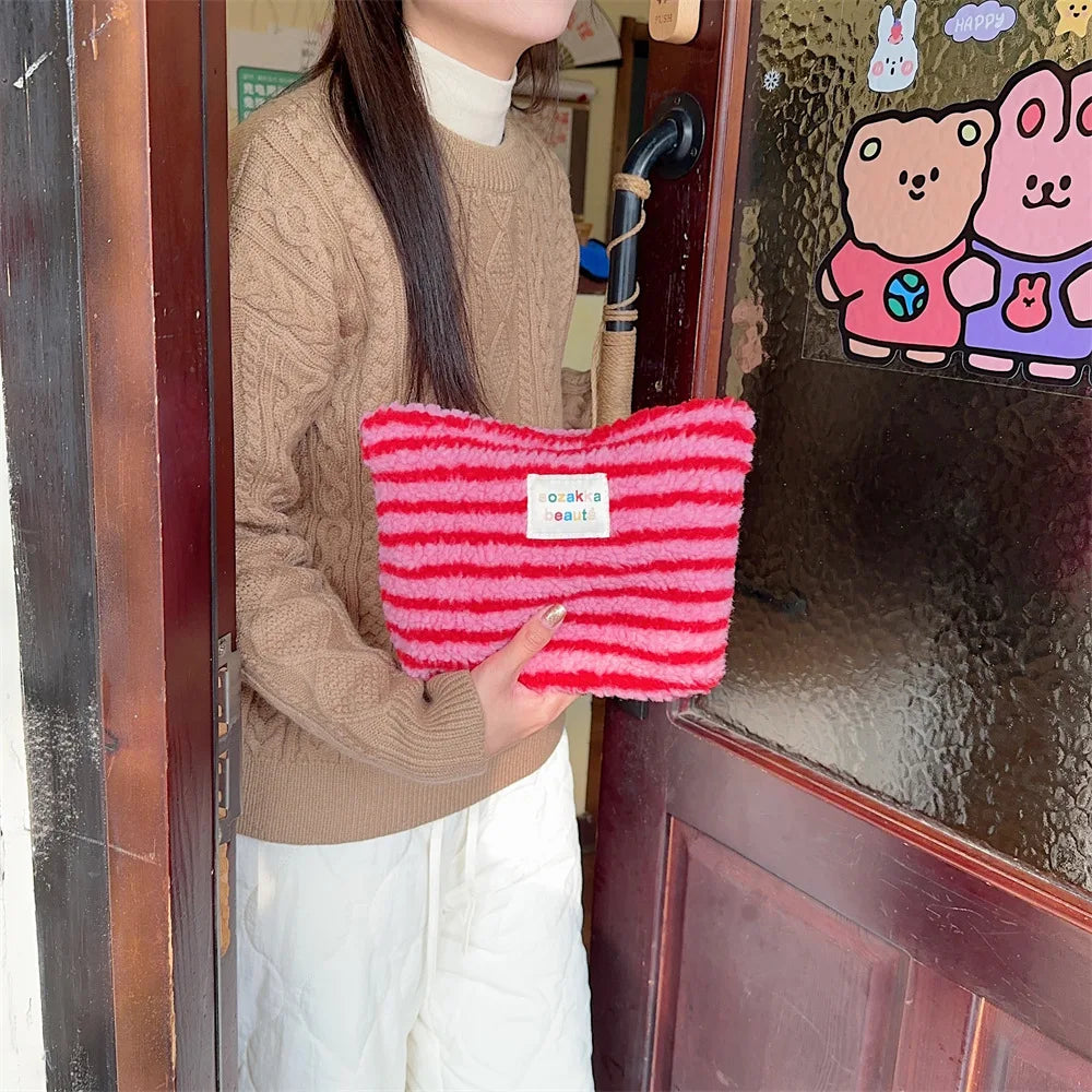 Donna con maglione marrone tiene una borsa a righe rosse e rosa, davanti a una porta con adesivi. Accessorio moda invernale, stile casual.