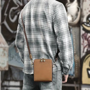 Uomo con camicia a quadri e jeans indossa borsa a tracolla in pelle marrone, stile casual urbano. Accessorio moda maschile, street style.