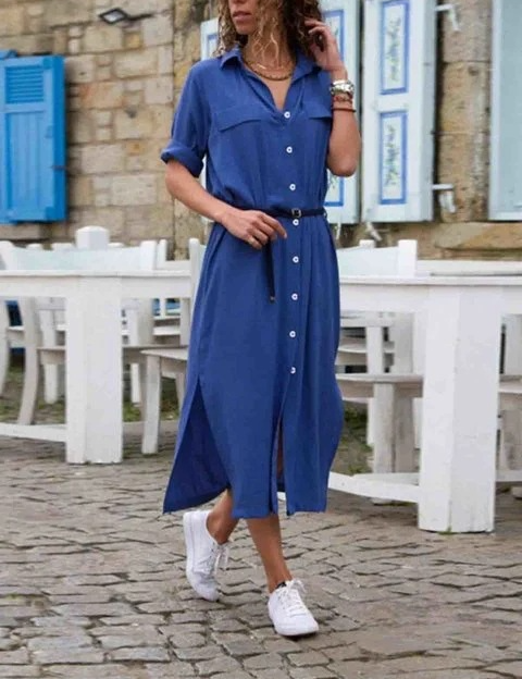 Donna con abito camicia blu, cintura nera, scarpe da ginnastica bianche, cammina su strada acciottolata. Moda casual estiva, look urbano elegante.