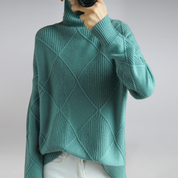 Maglione verde acqua a collo alto con motivo a rombi, indossato da una persona, abbinato a pantaloni bianchi. Moda autunnale elegante e casual.