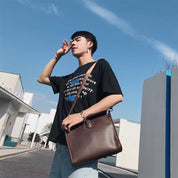 Giovane uomo con borsa a tracolla in pelle marrone, indossa t-shirt nera con scritte e jeans chiari, sfondo urbano e cielo azzurro.