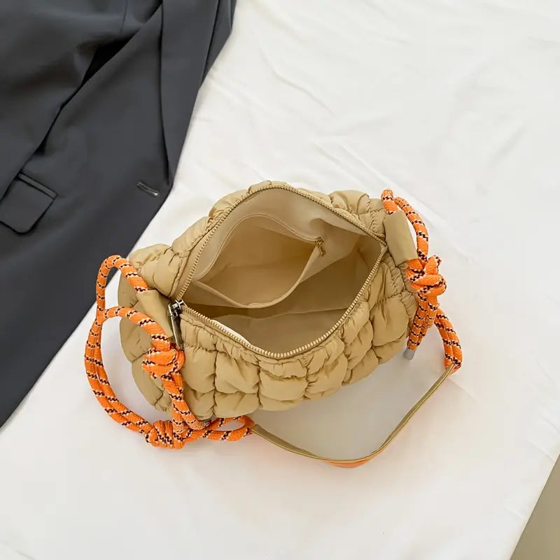 Borsa trapuntata beige con manici a corda arancioni, aperta su un tavolo bianco accanto a una giacca grigia. Design moderno e spazioso.