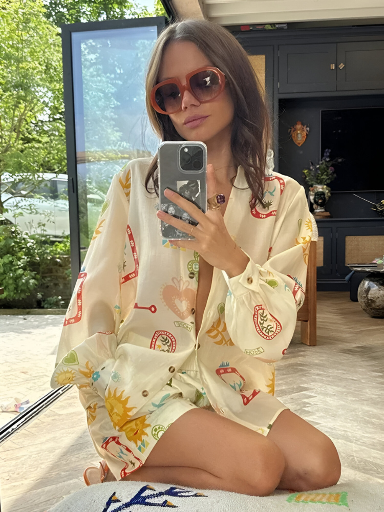 Donna con occhiali da sole e camicia colorata, scatta un selfie con smartphone in una stanza luminosa. Moda estiva, stile casual, interni moderni.