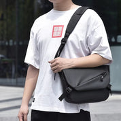 Uomo con borsa a tracolla nera in pelle, indossa maglietta bianca. Accessorio moda maschile, casual, urbano, perfetto per lavoro o viaggio.