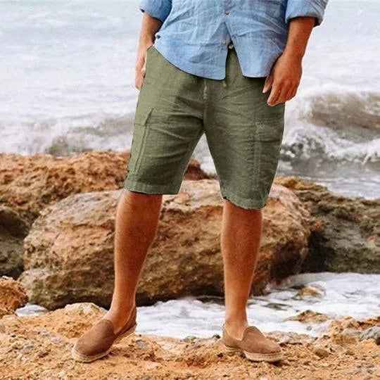 Uomo in pantaloncini verdi e camicia azzurra su spiaggia rocciosa, indossa mocassini marroni. Moda estiva casual, abbigliamento maschile mare.