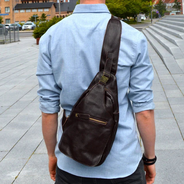 Uomo con zaino a tracolla in pelle marrone, indossa camicia azzurra. Accessorio moda maschile, stile urbano, pratico per viaggi e lavoro.