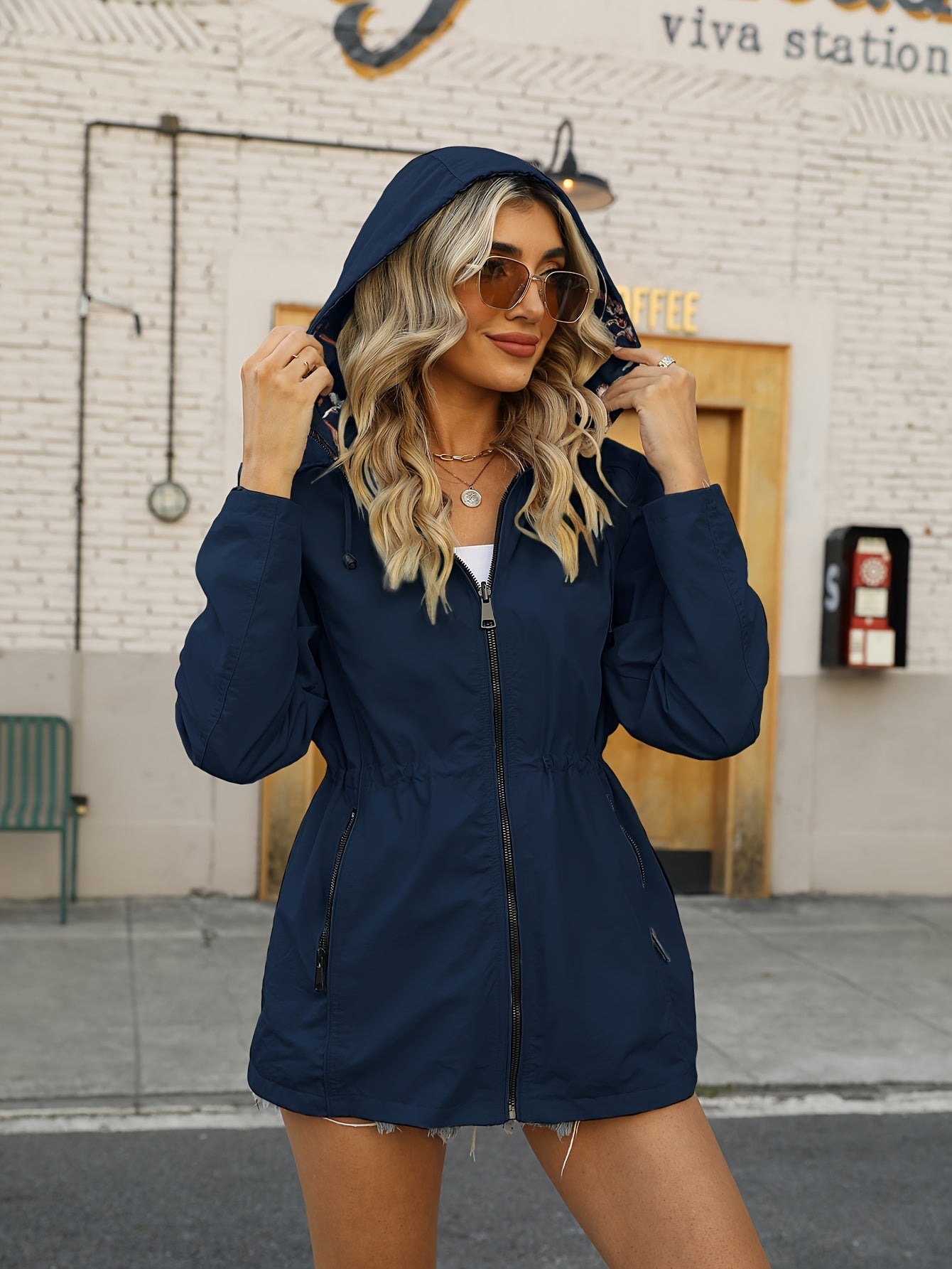 Donna con giacca a vento blu, cappuccio alzato, occhiali da sole, davanti a un edificio urbano. Moda autunnale, abbigliamento casual donna.