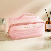Borsa da toilette rosa trasparente con cerniera, manico superiore, accanto a flacone spray e diffusore. Ideale per viaggi e organizzazione cosmetici.