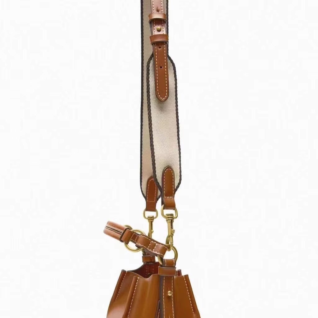 Borsa a secchiello in pelle marrone con tracolla regolabile in tessuto beige, dettagli in metallo dorato. Accessorio moda elegante e versatile.