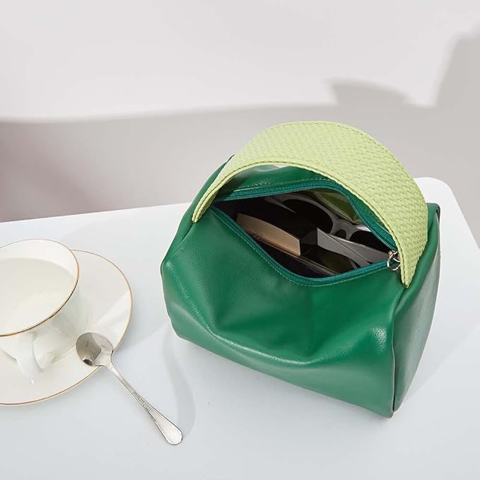 Borsa verde in pelle con manico intrecciato giallo, aperta su tavolo bianco. Include tazza da tè e cucchiaio. Accessorio moda elegante e moderno.