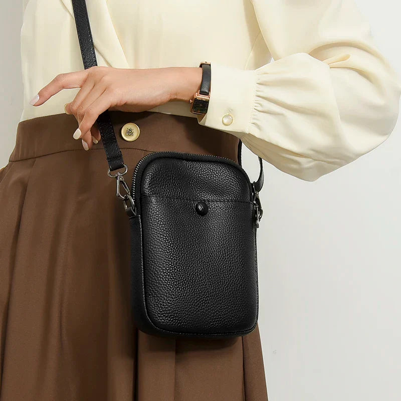 Borsa a tracolla in pelle nera, elegante e compatta, indossata da una donna con camicia bianca e gonna marrone. Accessorio moda femminile.