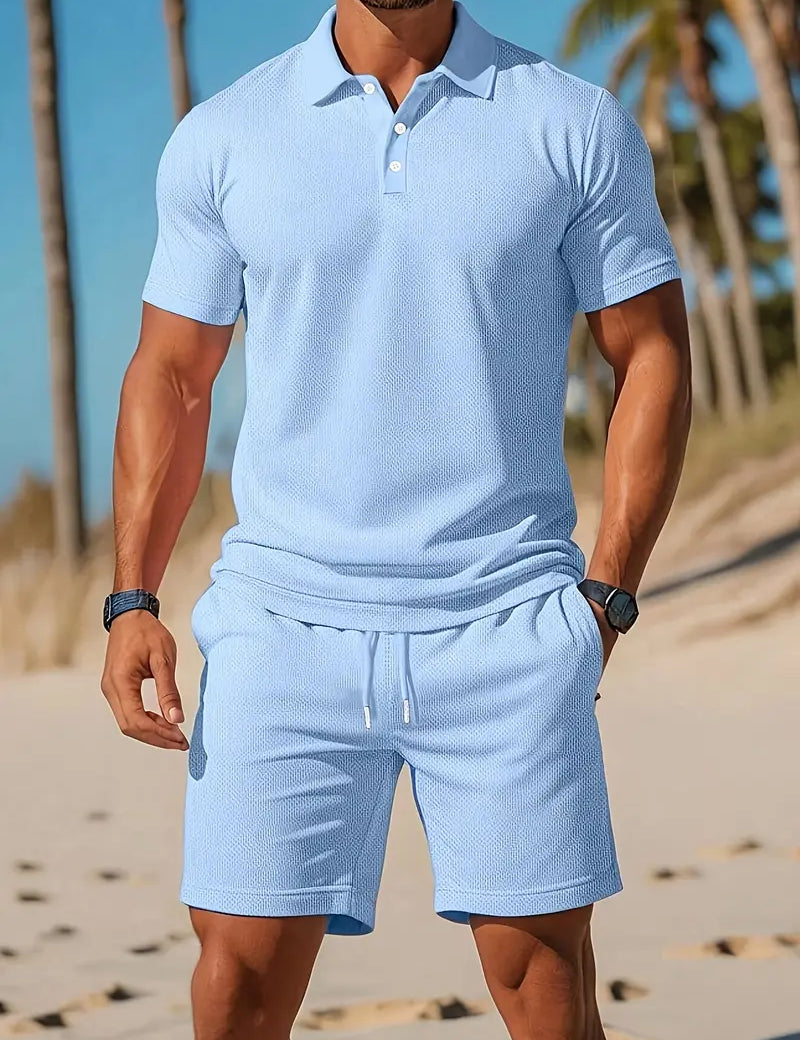 Uomo in spiaggia con polo azzurra e pantaloncini coordinati, palme sullo sfondo. Abbigliamento estivo casual, moda uomo, look vacanza.