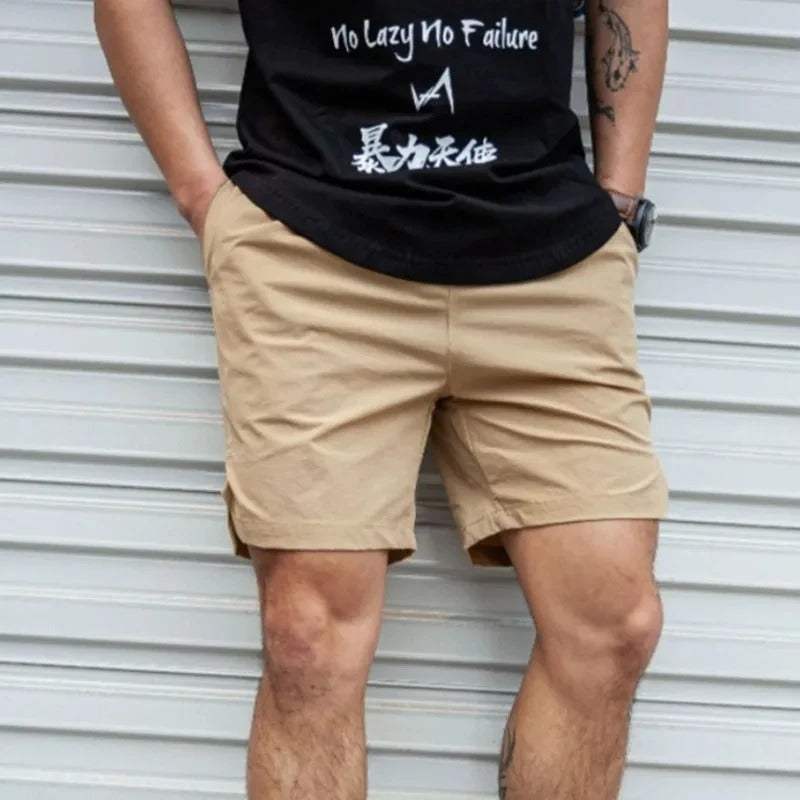Uomo in pantaloncini beige e maglietta nera con scritte bianche, posa casual davanti a una porta metallica. Moda estiva, abbigliamento casual uomo.