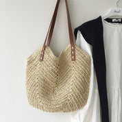 Borsa in paglia intrecciata con manici in pelle marrone, appesa accanto a un abito bianco. Accessorio estivo elegante e alla moda.