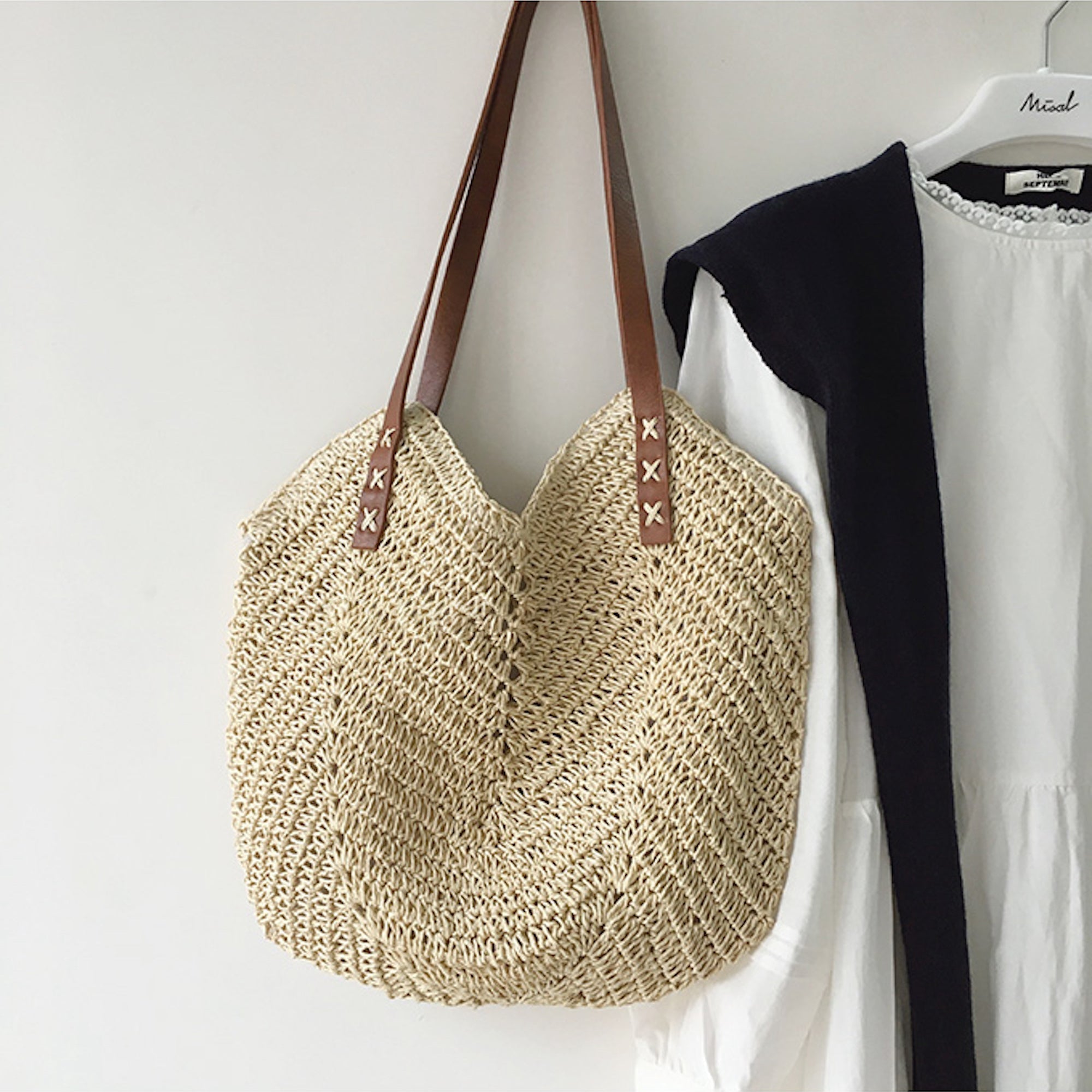 Borsa in paglia intrecciata con manici in pelle marrone, appesa accanto a un abito bianco. Accessorio estivo elegante e alla moda.