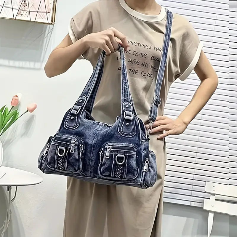 Borsa a tracolla in denim blu con tasche multiple e dettagli in metallo, indossata da una persona in abito beige. Accessorio moda casual e versatile.