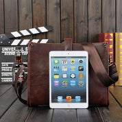 Borsa in pelle marrone con tablet iPad, clapperboard e bottiglia su sfondo di legno. Accessori eleganti per lavoro e tempo libero.