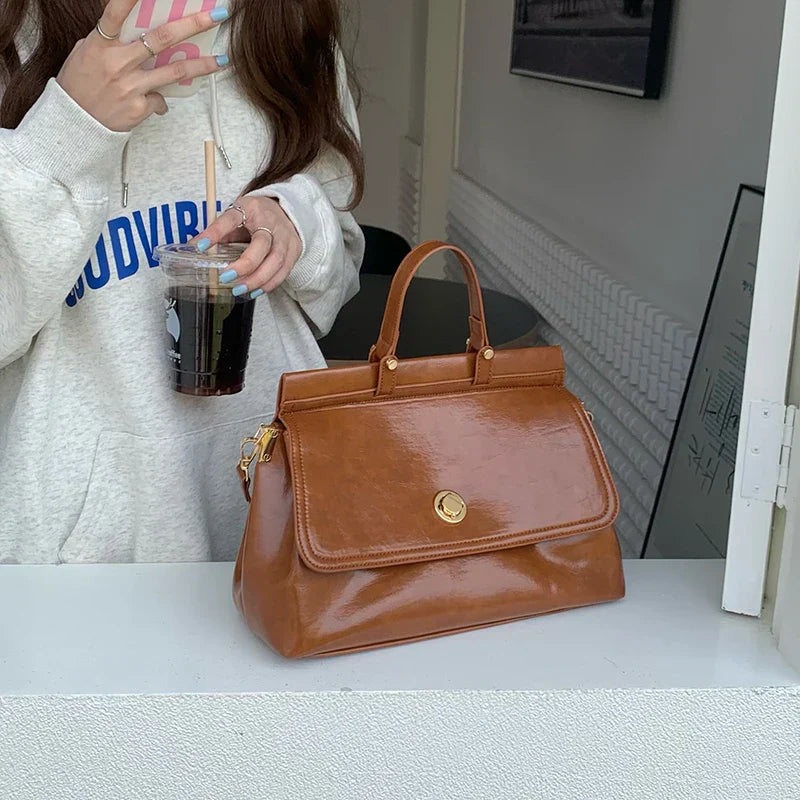 Borsa a mano in pelle marrone lucida su tavolo bianco, con chiusura a bottone dorato. Donna con felpa bianca e caffè freddo sullo sfondo.