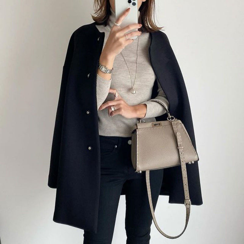Donna con cappotto nero, maglione grigio, borsa a tracolla beige e smartphone. Moda autunnale elegante, abbigliamento casual chic.