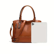Borsa a mano in pelle marrone con tracolla regolabile, dettagli in metallo, accanto a un tablet bianco. Elegante accessorio moda donna.