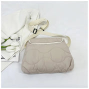 Borsa a tracolla imbottita beige su sfondo bianco, con cinturino regolabile e fiori bianchi accanto. Design moderno, ideale per moda casual.