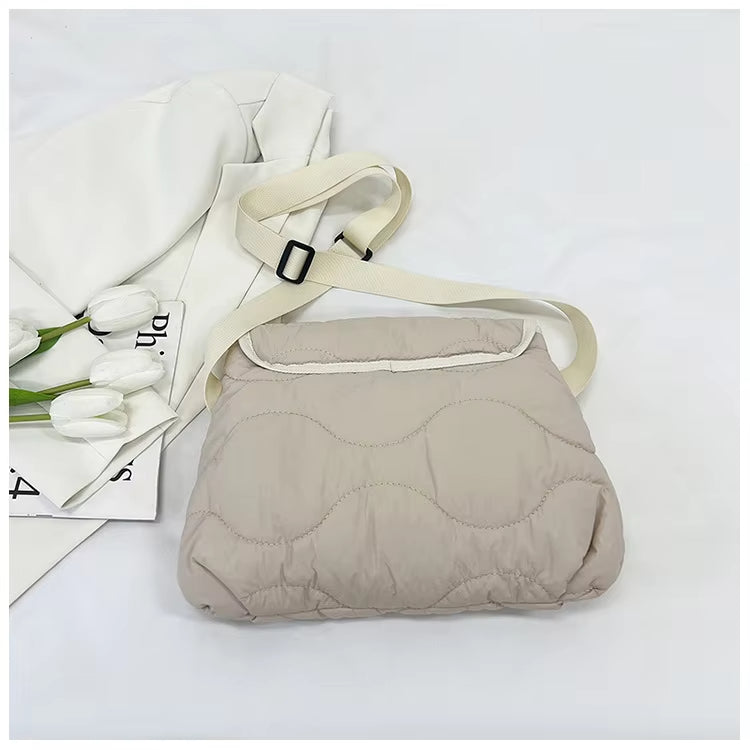 Borsa a tracolla imbottita beige su sfondo bianco, con cinturino regolabile e fiori bianchi accanto. Design moderno, ideale per moda casual.