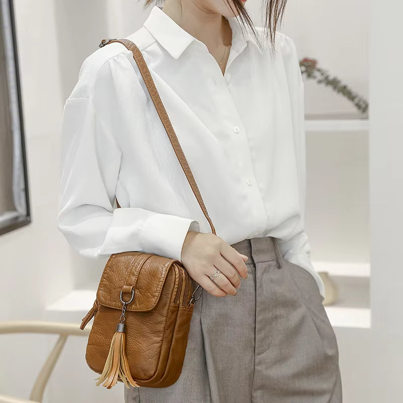 Donna con camicia bianca e pantaloni grigi indossa borsa a tracolla in pelle marrone con nappina. Elegante accessorio moda femminile.