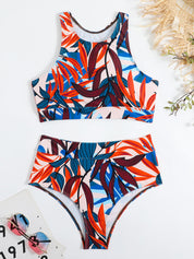 Costume da bagno donna a due pezzi con stampa tropicale colorata, top sportivo e slip a vita alta, occhiali da sole e cappello di paglia sullo sfondo.