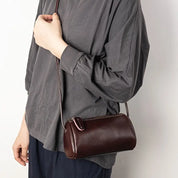 Borsa a tracolla cilindrica in pelle marrone scuro, indossata da una persona con maglia grigia. Accessorio moda elegante e versatile.