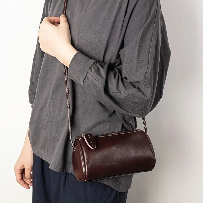 Borsa a tracolla cilindrica in pelle marrone scuro, indossata da una persona con maglia grigia. Accessorio moda elegante e versatile.