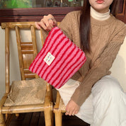 Donna con maglione marrone tiene borsa a righe rosse e bianche in pelliccia sintetica, seduta su sedia di bambù. Accessorio moda autunno inverno.