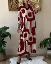 Abito lungo donna, fantasia geometrica rosso e beige, maniche lunghe, stile casual, su tappeto bianco, piante decorative ai lati.