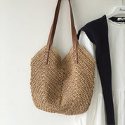 Borsa a tracolla in paglia intrecciata con manici in pelle marrone, appesa accanto a un abito bianco. Accessorio estivo elegante e casual.