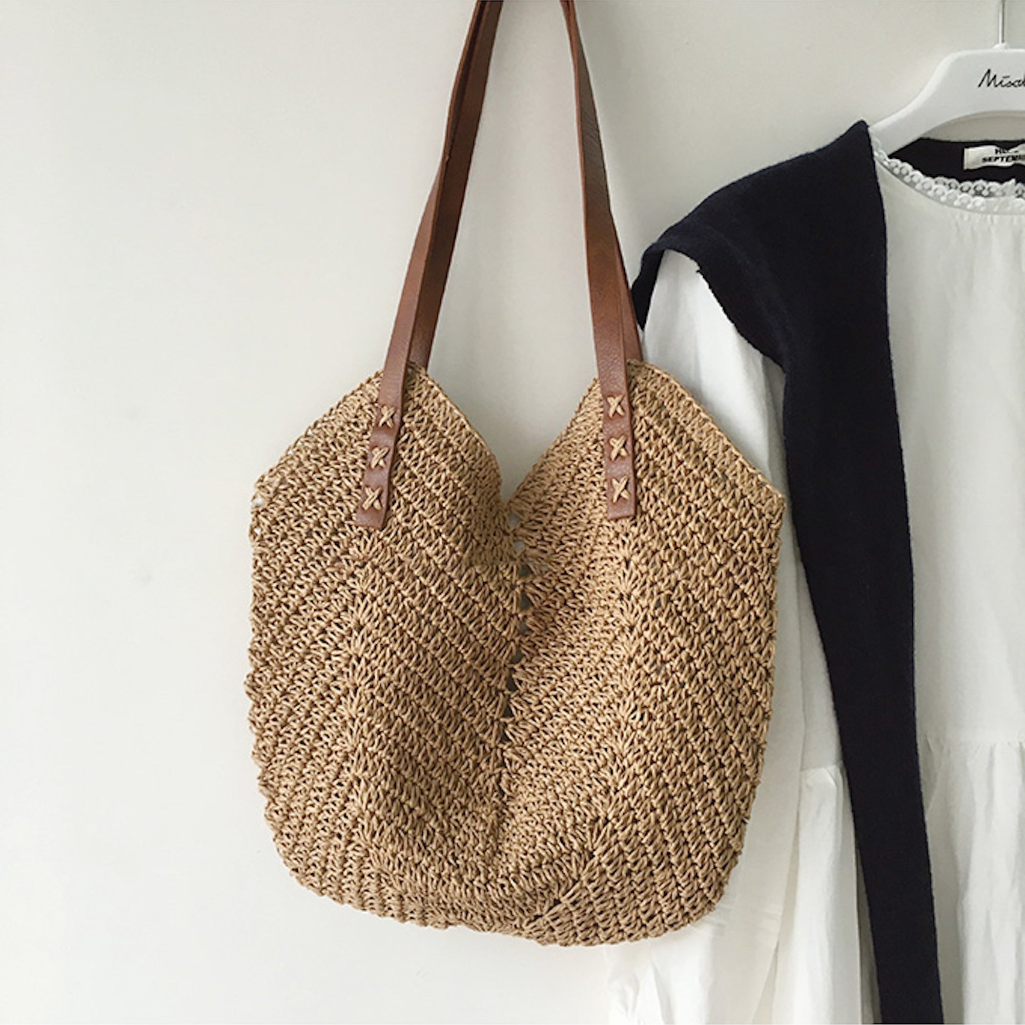 Borsa a tracolla in paglia intrecciata con manici in pelle marrone, appesa accanto a un abito bianco. Accessorio estivo elegante e casual.