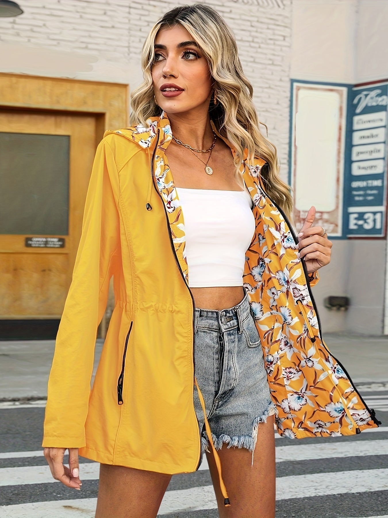 Giacca impermeabile gialla da donna con fodera floreale, indossata su top bianco e shorts di jeans. Moda casual estiva, stile urbano.