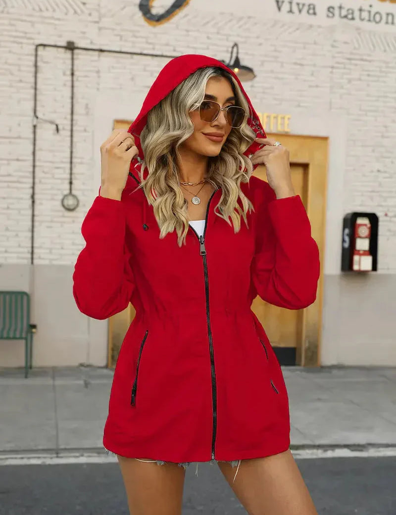 Donna con giacca rossa con cappuccio, occhiali da sole, davanti a un edificio urbano. Moda autunnale, abbigliamento casual, stile urbano.