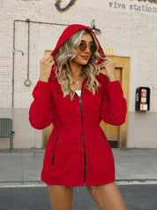 Donna con giacca rossa con cappuccio, occhiali da sole, davanti a un edificio urbano. Moda autunnale, abbigliamento casual, stile urbano.