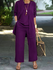 Donna in elegante completo viola, pantaloni larghi e giacca lunga, con borsa a tracolla marrone e sandali con zeppa. Look moda autunnale chic.