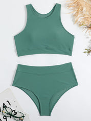 Costume da bagno verde a due pezzi, top sportivo e slip a vita alta, su sfondo bianco con occhiali e accessori estivi. Moda mare donna.
