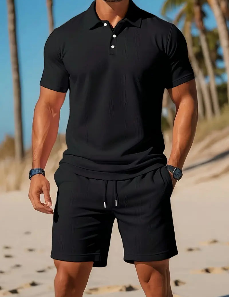 Uomo in spiaggia con polo nera e pantaloncini coordinati, palme sullo sfondo. Moda estiva maschile, abbigliamento casual elegante.