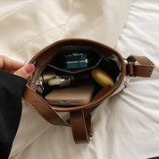 Interno di una borsa a tracolla marrone su letto bianco, contenente smartphone, rossetto, profumo e portafoglio. Accessori moda donna eleganti.