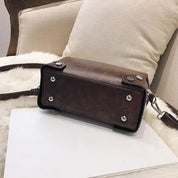 Borsa a tracolla in pelle marrone su sedia beige, design rettangolare con dettagli metallici, ideale per moda donna e accessori eleganti.